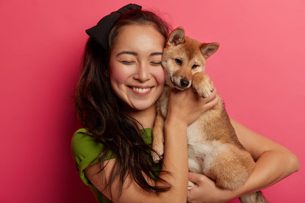 Happy woman pet owner holds cute dog near face, carries shiba inu puppy, smiles pleasantly, enjoys sweet moment, isolated over pink background. Friendship between human and animal. Best friends